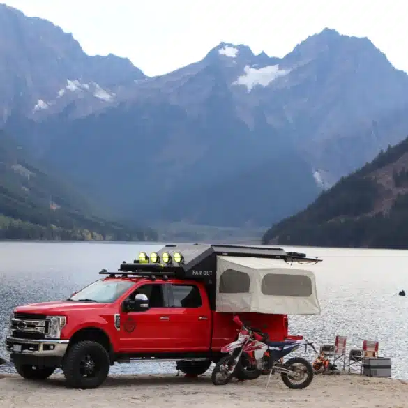 Roter Pick-up mit Far Out Canopy Truck Camper und zwei Motorrädern am Seeufer vor Bergkulisse