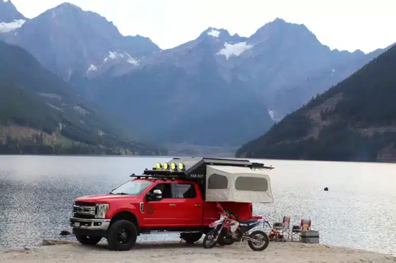 Roter Pick-up mit Far Out Canopy Truck Camper und zwei Motorrädern am Seeufer vor Bergkulisse