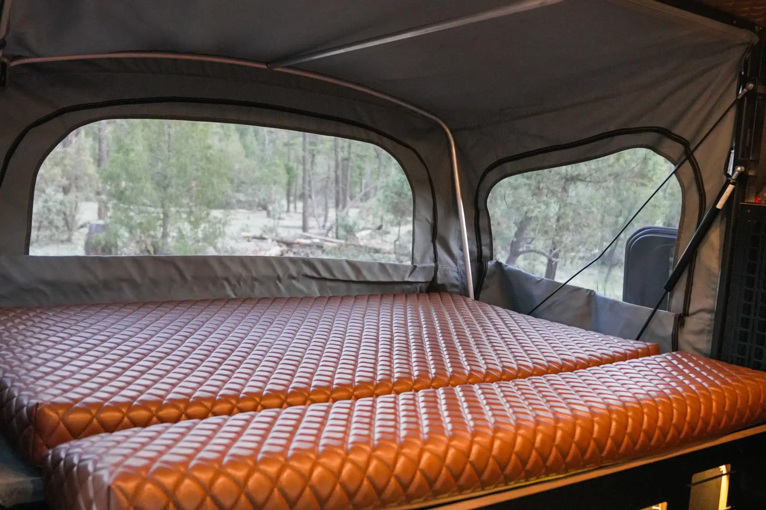 Schlafbereich mit brauner, gesteppter Matratze im Far Out Canopy Truck Camper, Blick nach draußen durch große Fenster mit Moskitonetz
