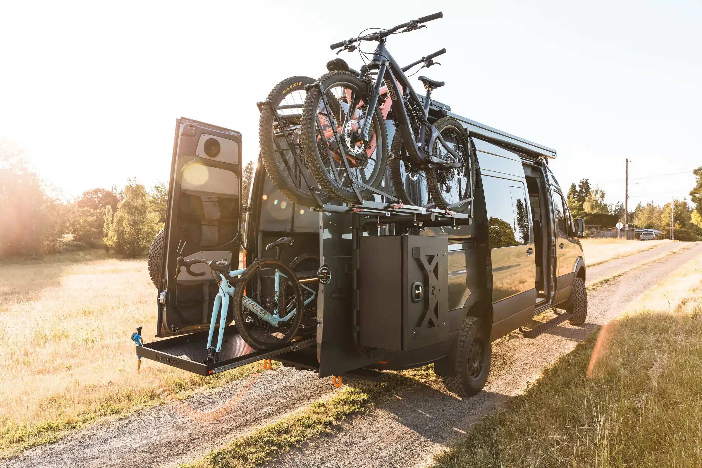 Mercedes Sprinter 4x4 Campervan mit geöffneten Hecktüren, Fahrradträger und Outdoor-Ausrüstung auf einem Feldweg