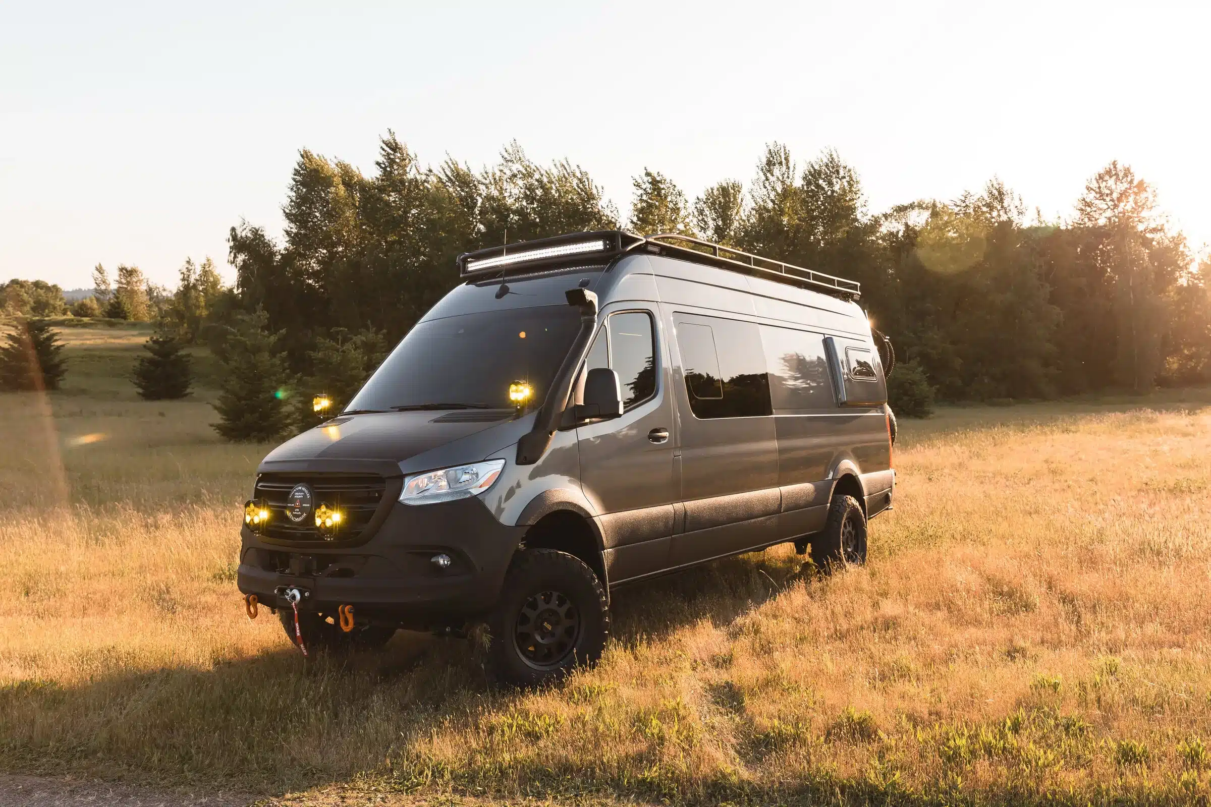 Gus 4x4 Sprinter Campervan mit Offroad-Umbau, LED-Lichtleisten und Dachträger auf einer Wiese bei Sonnenuntergang