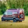 Gwanang Campervan auf Mercedes-Benz Sprinter 170 4x4, burgunderrot, mit Offroad-Reifen, Dachträger und Reserverad, steht auf einer Wiese vor Wald