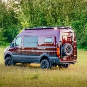 Gwanang Campervan auf Mercedes-Benz Sprinter 170 4x4, burgunderrot, mit Offroad-Reifen, Dachträger und Reserverad, steht auf einer Wiese vor Wald