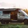 Seitenansicht eines ausgebauten HIDEOUT Sprinter 170 4x4 Vans mit geöffnetem Schiebetürbereich, Markise und Offroad-Ausstattung im Sonnenuntergang.