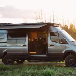 Seitenansicht eines ausgebauten HIDEOUT Sprinter 170 4x4 Vans mit geöffnetem Schiebetürbereich, Markise und Offroad-Ausstattung im Sonnenuntergang.