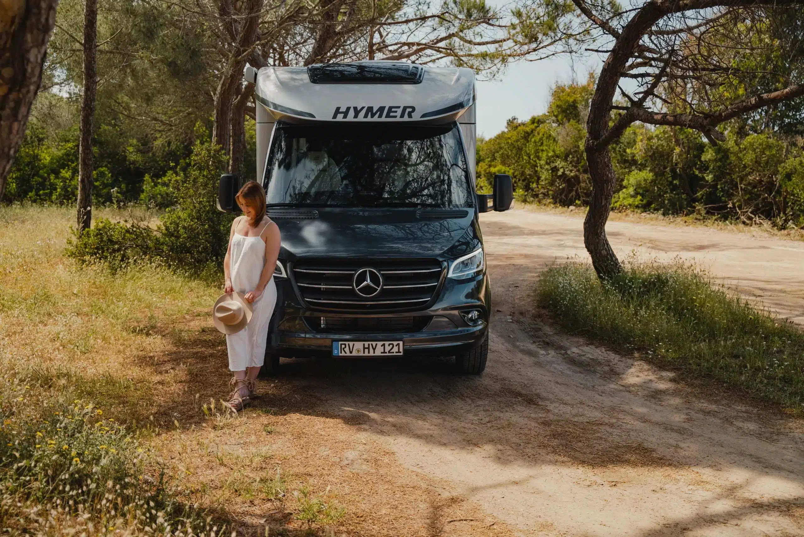 HYMER B-Klasse ModernComfort T Wohnmobil steht auf einem naturbelassenen Weg, eine Frau in Sommerkleidung steht davor.