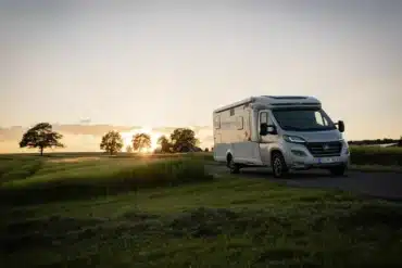 HYMER Exsis-t Pure Wohnmobil fährt bei Sonnenuntergang über eine Landstraße, umgeben von Feldern und Bäumen