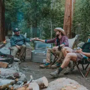 Drei Camper sitzen am Lagerfeuer im Wald, neben ihnen steht eine geöffnete ICECO APL35 Kompressor-Kühlbox