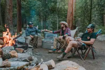 Drei Camper sitzen am Lagerfeuer im Wald, neben ihnen steht eine geöffnete ICECO APL35 Kompressor-Kühlbox