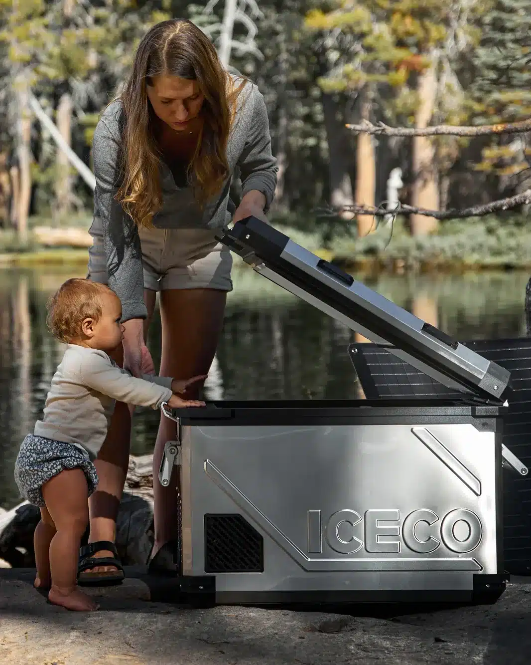 Frau und Kleinkind nutzen eine silberne ICECO APL55 Kompressor-Kühlbox am Seeufer im Wald