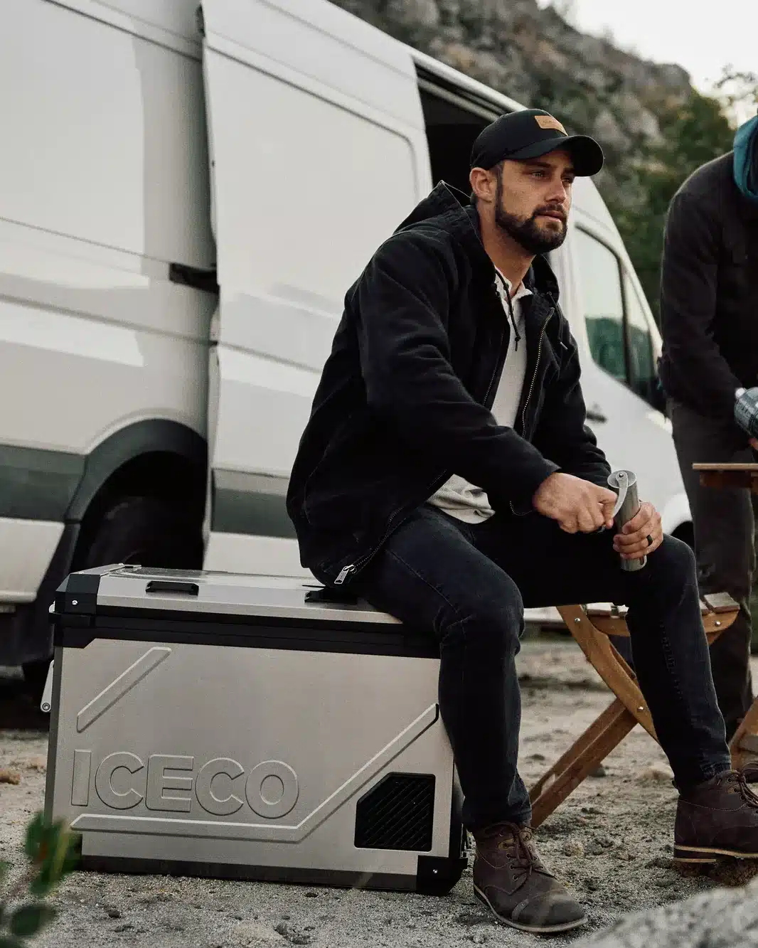 Mann sitzt auf einer silbernen ICECO APL55 Kompressor-Kühlbox vor einem Campervan in natürlicher Umgebung