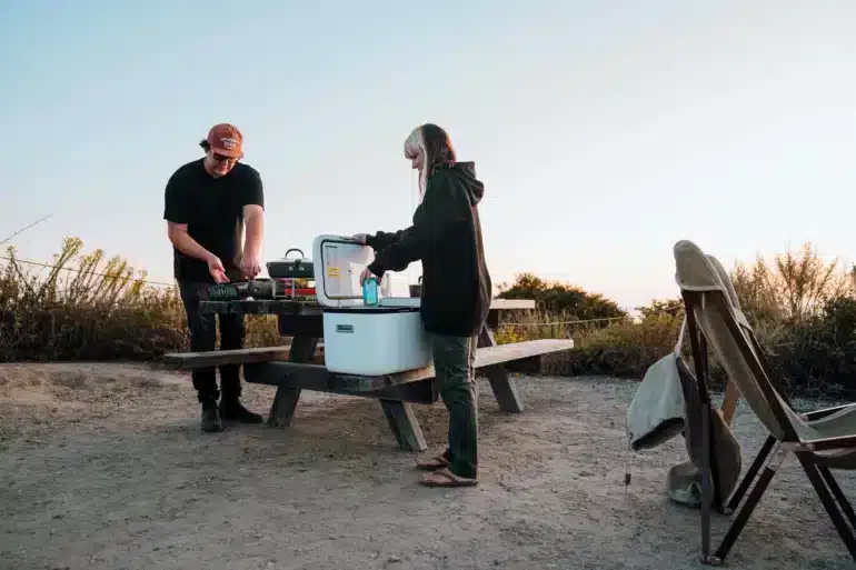 Zwei Personen am Picknicktisch mit geöffneter ICECO GO20 Kompressor-Kühlbox Dual Zone beim Camping