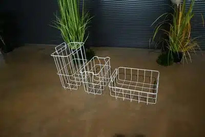 Drei weiße Drahtkörbe unterschiedlicher Größe stehen auf einem Betonboden, im Hintergrund sind grüne Pflanzen zu sehen.