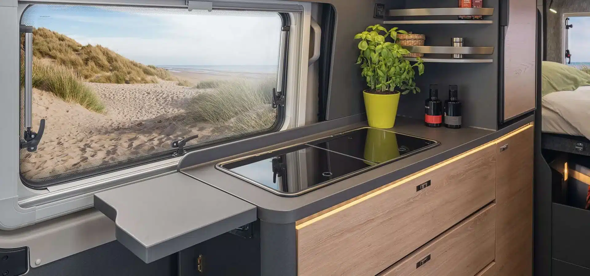 Küchenzeile im KNAUS BOXLIFE Camper Van mit Induktionskochfeld, Kräutertopf und Blick durch das Fenster auf eine Dünenlandschaft