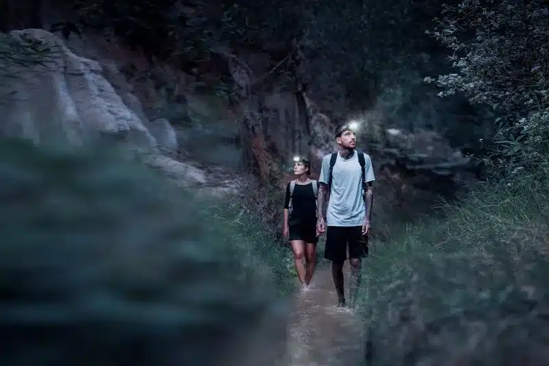 Zwei Menschen mit Ledlenser Stirnlampen beim nächtlichen Wandern durch einen Wasserlauf im Grünen.
