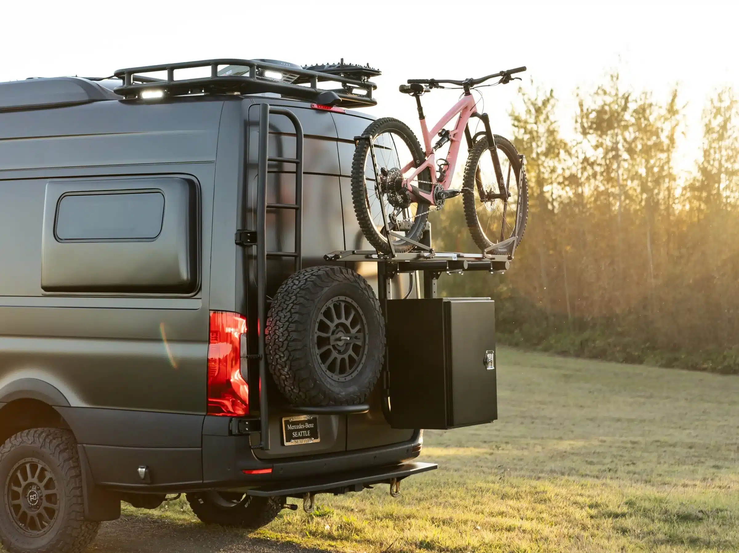 Heckansicht eines ausgebauten Mercedes Sprinter Vans mit montiertem Fahrradträger, Reserverad und Staubox im Sonnenuntergang