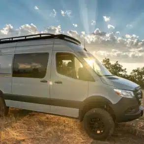 Mercedes Sprinter 4x4 Azure Campervan mit Dachträger und Offroad-Bereifung auf einer Wiese bei Sonnenuntergang
