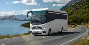 Der Morelo Empire Liner fährt auf einer kurvigen Landstraße entlang eines Sees mit Bergen im Hintergrund.