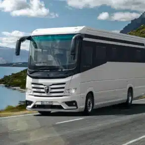 Der Morelo Empire Liner fährt auf einer kurvigen Landstraße entlang eines Sees mit Bergen im Hintergrund.