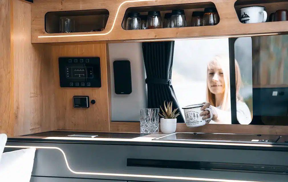 Blick auf die Küchenzeile und den Innenausbau eines Rhön Camp The Pace Campers mit Holzdekor, Gläsern, Tassen und einer Person am Fenster