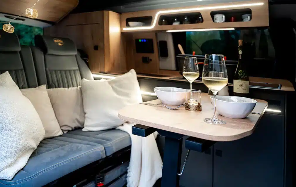 Innenansicht eines Campervans mit gemütlicher Sitzbank, Esstisch, zwei Gläsern Weißwein und moderner Küchenzeile