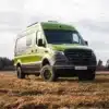 Grüner Rhön Camp THE SPIRIT Campervan auf Mercedes Sprinter Basis steht auf einer Wiese vor bewaldetem Hintergrund