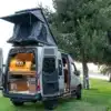Heckansicht eines hochwertigen Campervans mit geöffnetem Dachzelt und modernem Innenausbau, geparkt auf einer Wiese