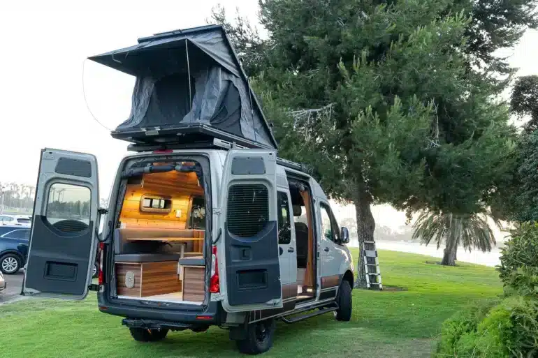 Heckansicht eines hochwertigen Campervans mit geöffnetem Dachzelt und modernem Innenausbau, geparkt auf einer Wiese