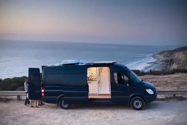Ein ausgebauter Mercedes Sprinter Campervan von Rogue Vans steht mit geöffneter Tür an einer Küste, der Innenraum mit Küche und Dusche ist sichtbar.