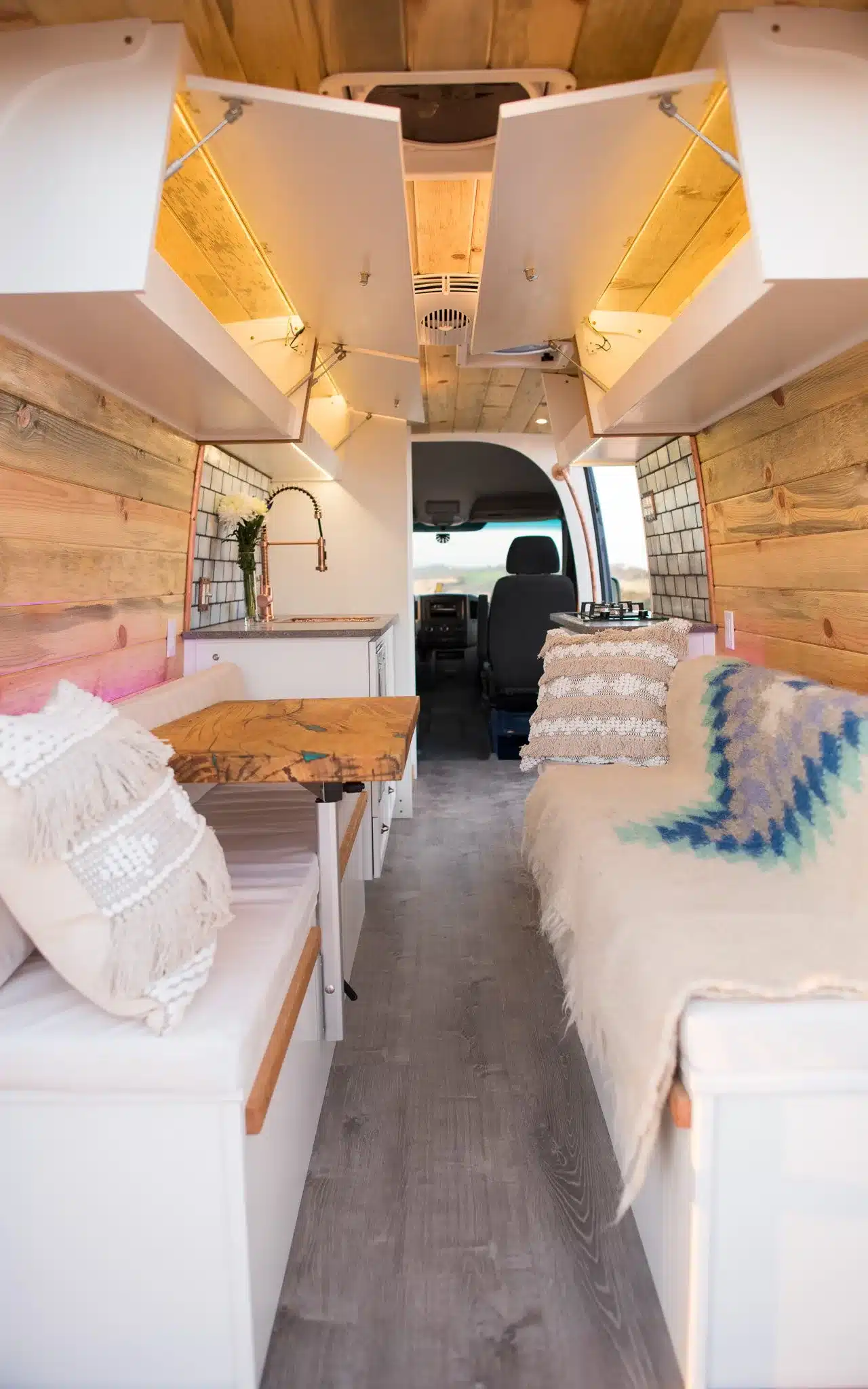 Blick in den Innenraum eines ausgebauten Campervans mit Holzverkleidung, Dinette, Küchenzeile und offenen Staufächern