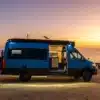 Blauer Rogue Vans Fox Den Campervan mit geöffneter Tür am Strand bei Sonnenuntergang, Innenraum sichtbar