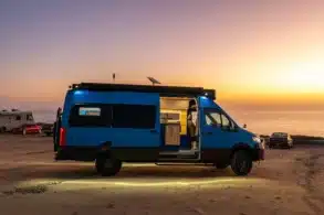 Blauer Rogue Vans Fox Den Campervan mit geöffneter Tür am Strand bei Sonnenuntergang, Innenraum sichtbar