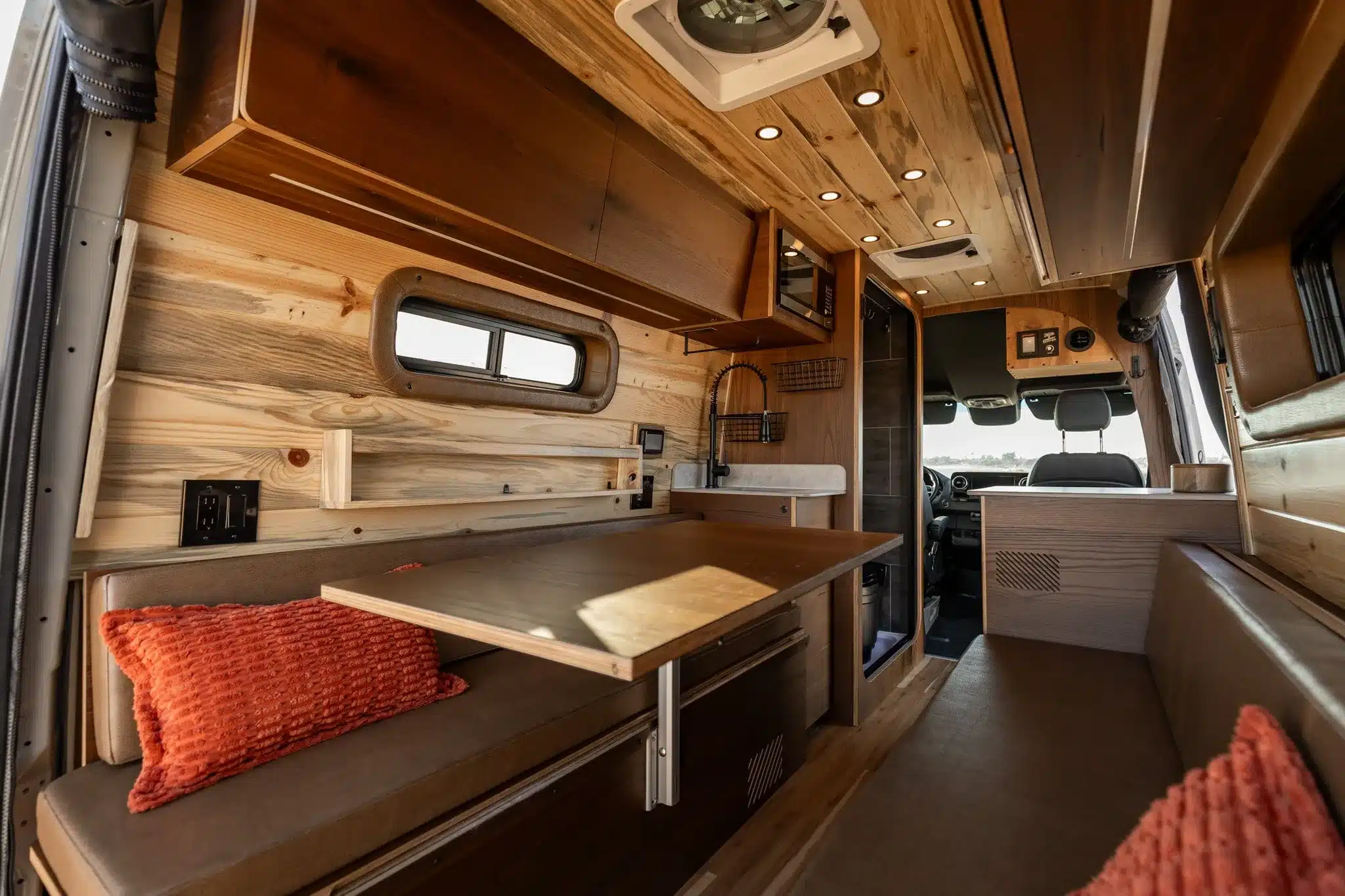Innenansicht eines ausgebauten Campervans mit Holzverkleidung, Sitzbänken, Tisch, Hängeschränken und Küchenzeile