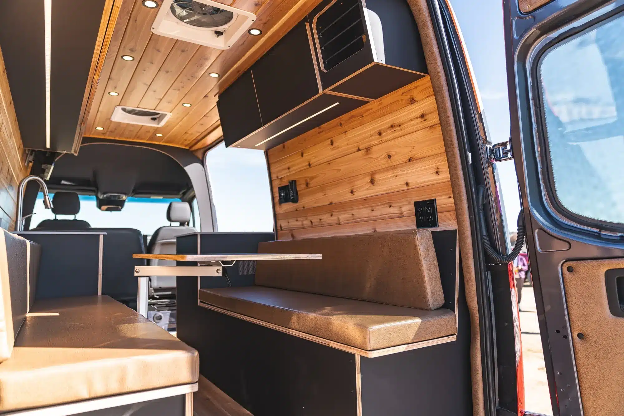 Innenansicht eines ausgebauten Campervans mit zwei gegenüberliegenden Sitzbänken, Tisch, Holzdecke und moderner Ausstattung