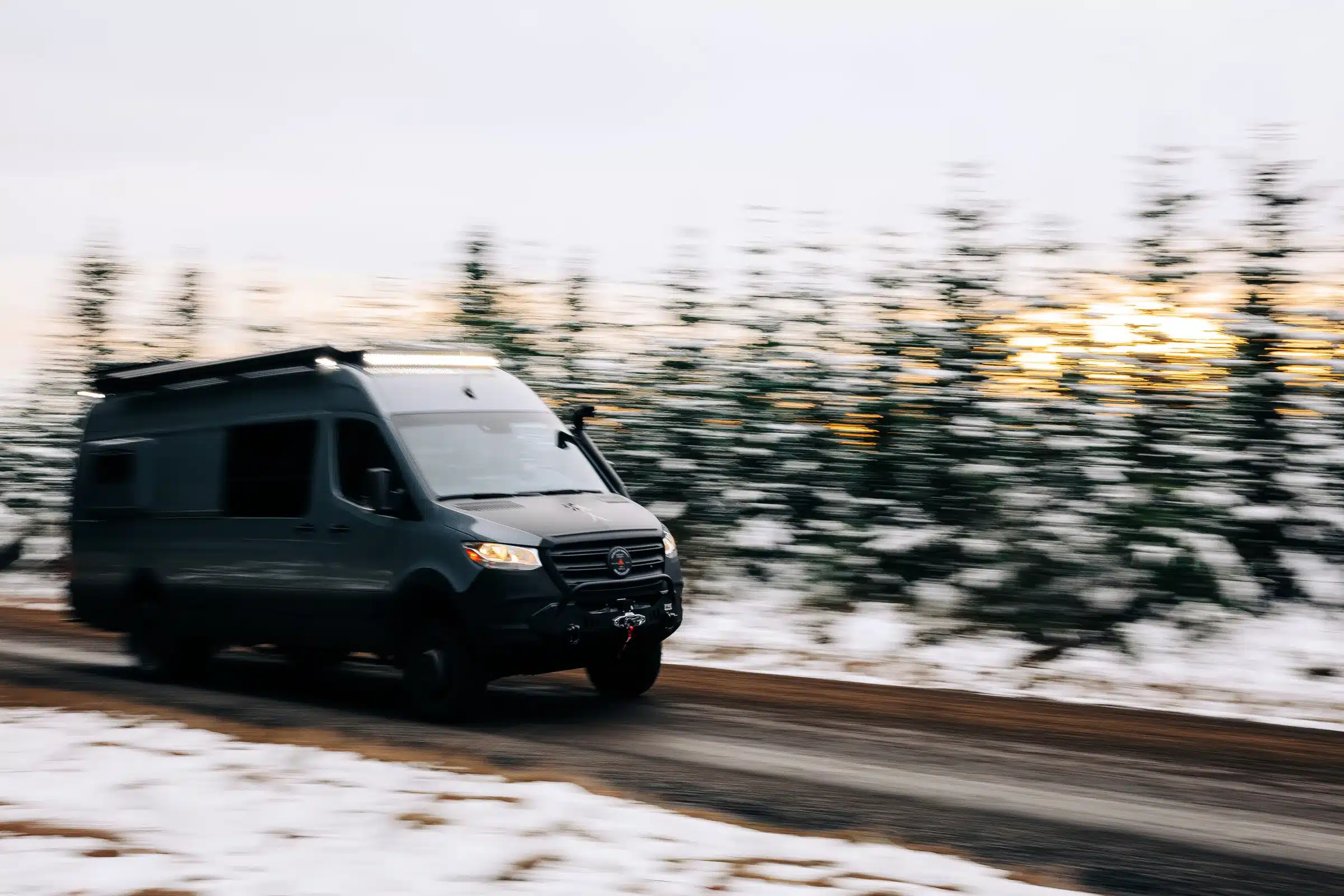 Ein schwarzer Mercedes Sprinter 170 EXT 4x4 Campervan fährt auf einer verschneiten Straße durch einen winterlichen Wald