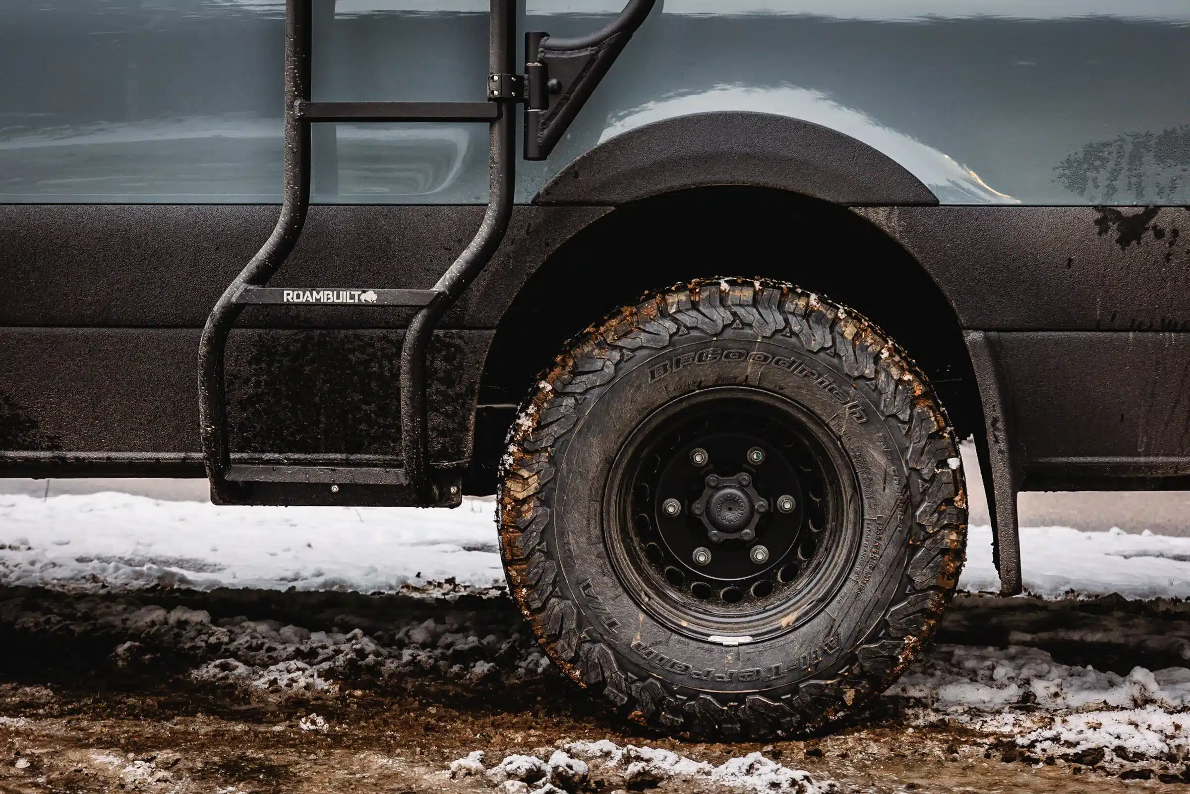 Nahaufnahme eines Offroad-Reifens mit BFGoodrich-Profil und Roambuilt-Leiter an einem Mercedes Sprinter 4x4 auf verschneitem Untergrund