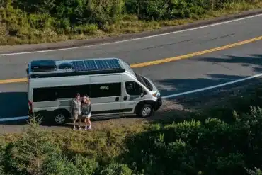 Safari Condo Ram ProMaster Go-Van 21 silberner Kastenwagen mit Solarpanel, geparkt am Straßenrand, zwei Personen davor, umgeben von Natur
