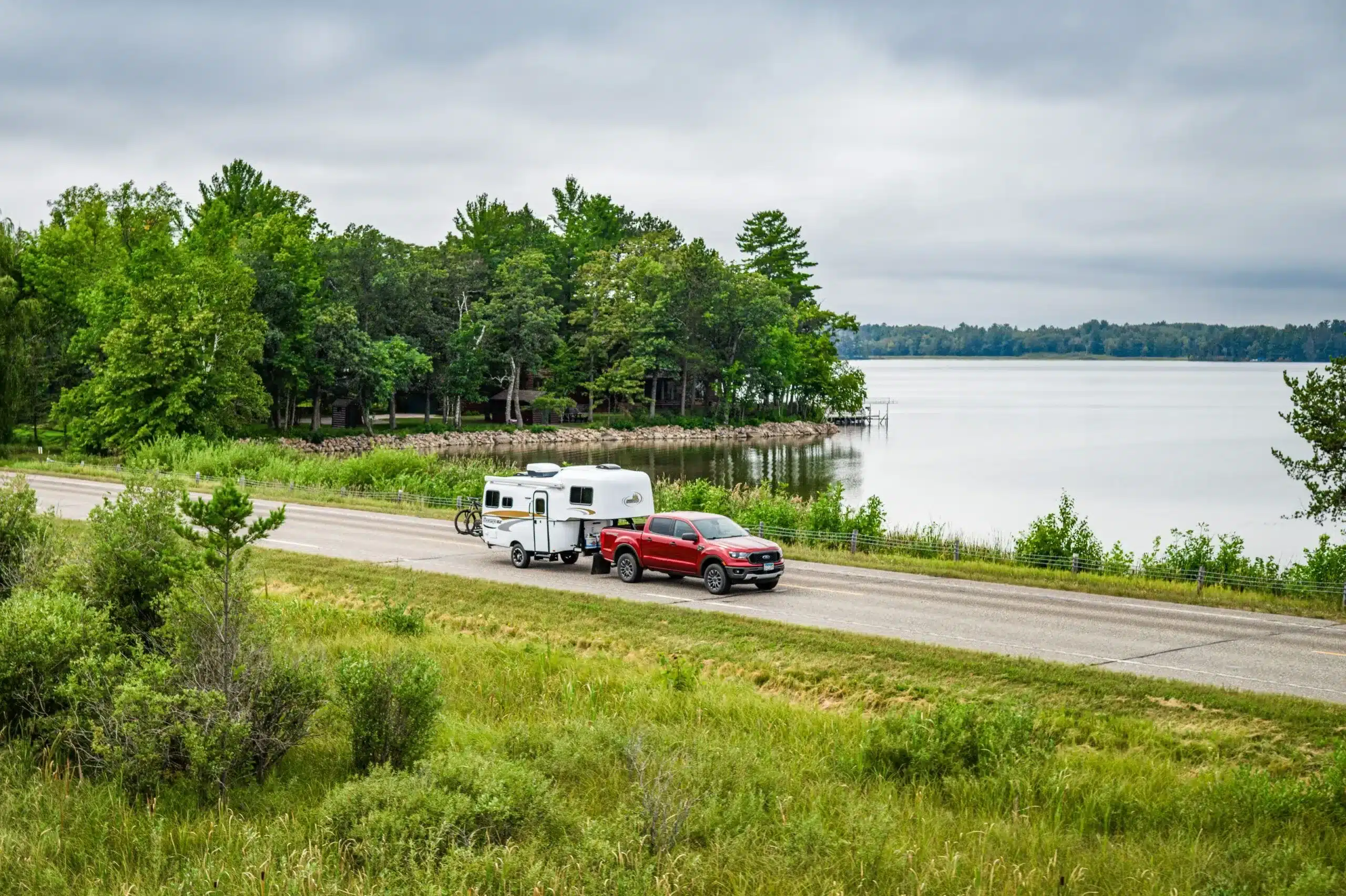 Ein roter Pickup zieht einen weißen Scamp 19' Trailer auf einer Landstraße entlang eines Sees, umgeben von grüner Natur und Bäumen.