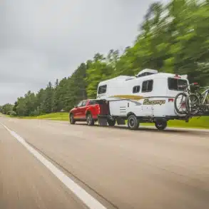 Ein roter Pickup zieht einen weißen Scamp 19' Trailer mit Fahrrädern auf einer Landstraße, umgeben von grüner Natur.