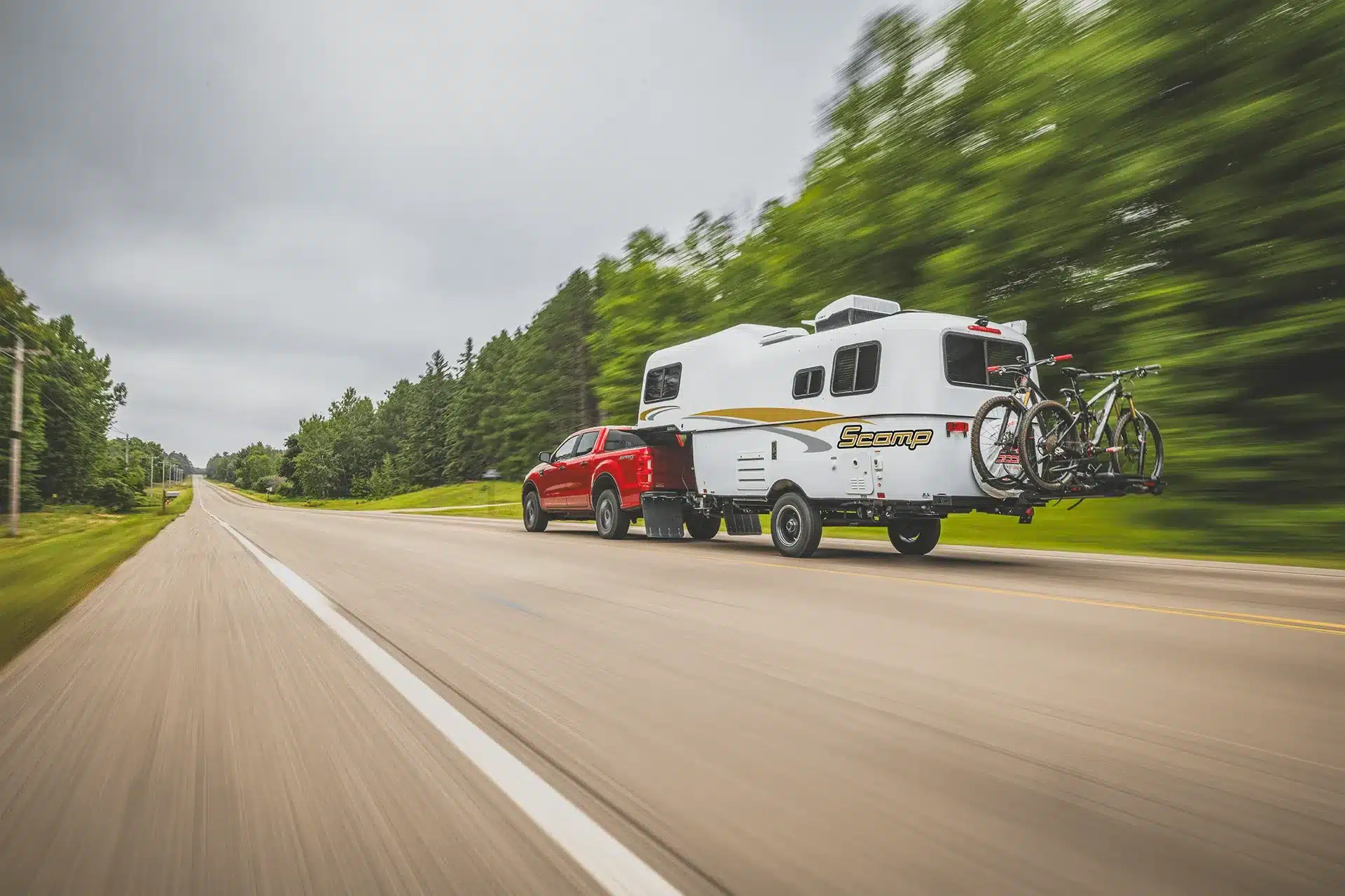 Ein roter Pickup zieht einen weißen Scamp 19' Trailer mit Fahrrädern auf einer Landstraße, umgeben von grüner Natur.