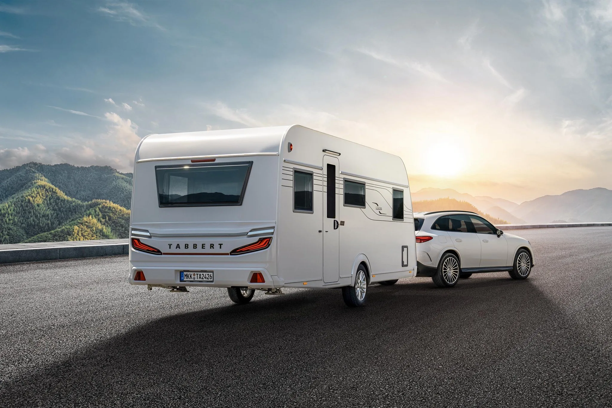 TABBERT Cazadora Wohnwagen wird von einem weißen Kombi auf einer Straße vor Bergkulisse gezogen