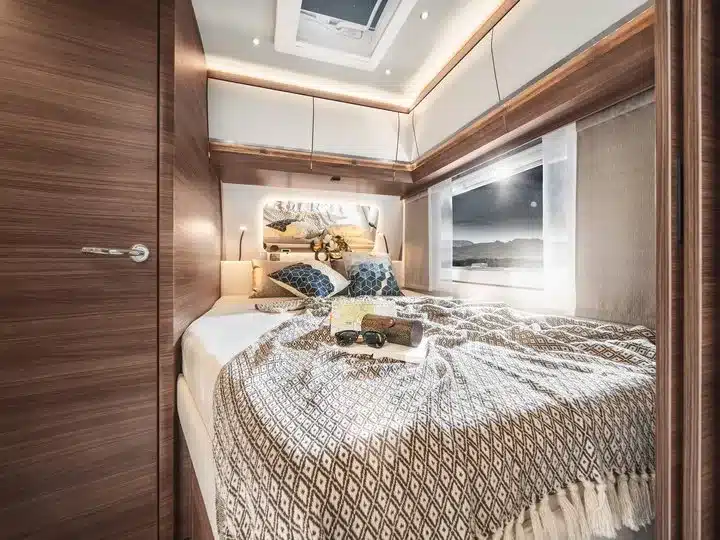 Hochwertig gestalteter Schlafbereich mit breitem Bett, stilvollen Kissen, Ober- und Unterschränken sowie großem Fenster im Tabbert Puccini Wohnwagen