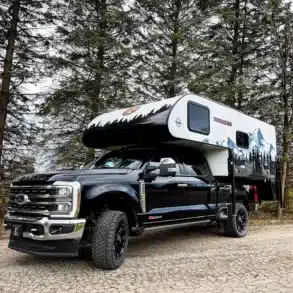 Ein TC14 Truck Camper von Modern Buggy RV montiert auf einem schwarzen Pick-up, geparkt auf einer Waldstraße, umgeben von Bäumen.