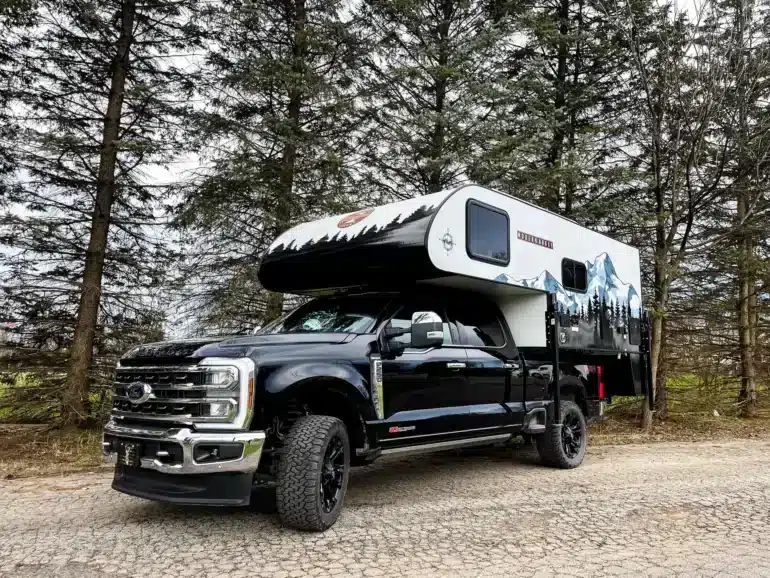 Ein TC14 Truck Camper von Modern Buggy RV montiert auf einem schwarzen Pick-up, geparkt auf einer Waldstraße, umgeben von Bäumen.