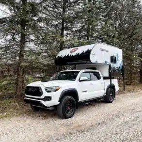 Weißer Pickup mit aufgesetztem Modern Buggy RV TC9 Truck Camper steht auf Waldweg