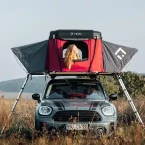 Frau sitzt im geöffneten TEDPop Hartschalen-Dachzelt auf einem Mini-Campervan, umgeben von Wiese und Natur