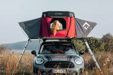 Frau sitzt im geöffneten TEDPop Hartschalen-Dachzelt auf einem Mini-Campervan, umgeben von Wiese und Natur