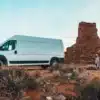 Weißer Hochdach-Campervan The Biggie steht in einer felsigen Wüstenlandschaft mit einer Person im Vordergrund