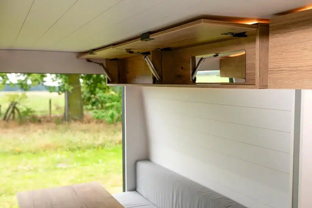 Offene Überkopf-Schränke aus Holz im Innenraum eines Campervans mit Blick auf Sitzbank und Tisch