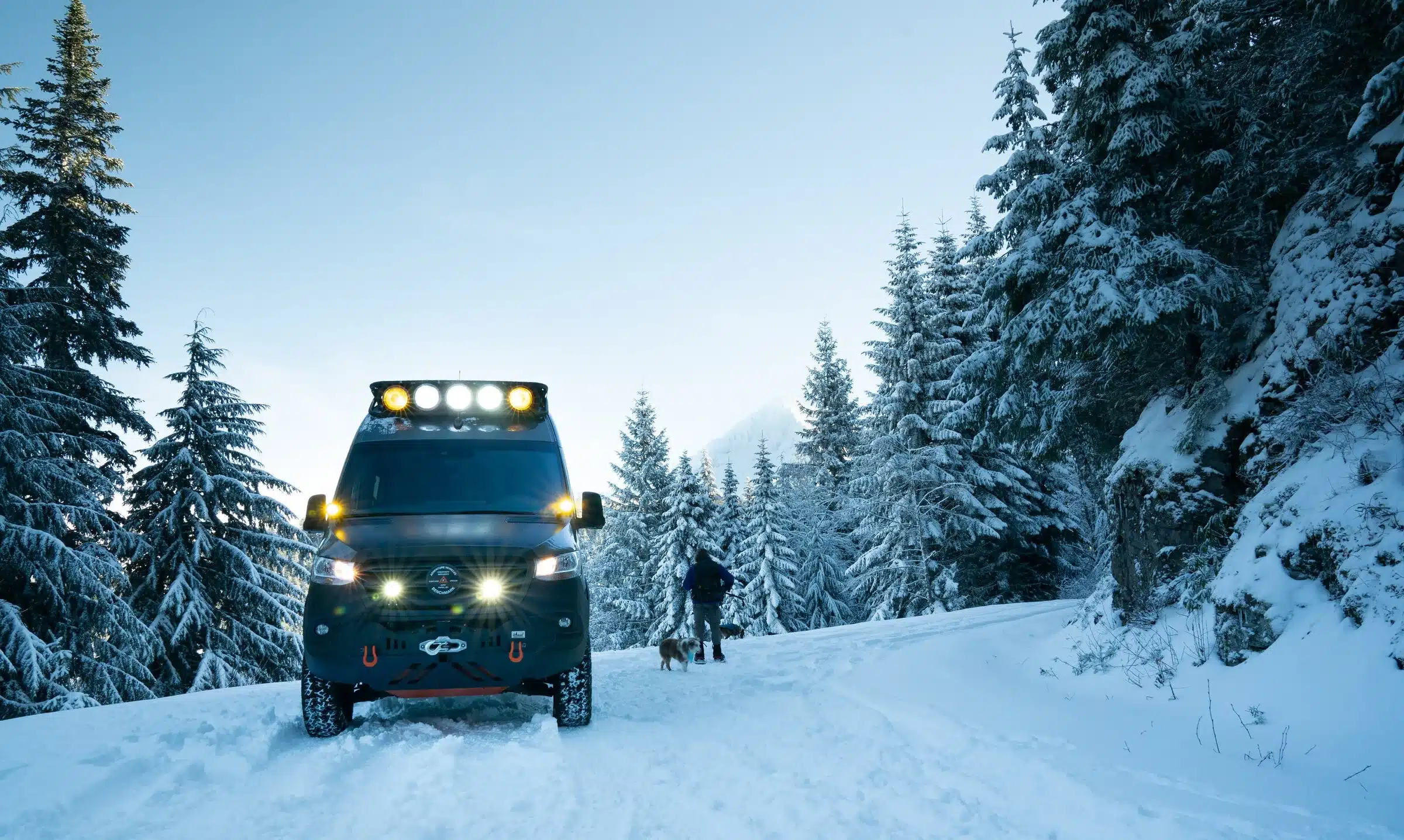 Mercedes Sprinter 170 4x4 Campervan mit Offroad-Ausstattung im verschneiten Wald, Person mit Hund im Hintergrund